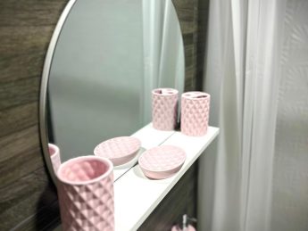 Bathroom with round mirror, white shelf, and pink accessories at Villa Sunrise, Flamingo Bay Villas Aruba.
